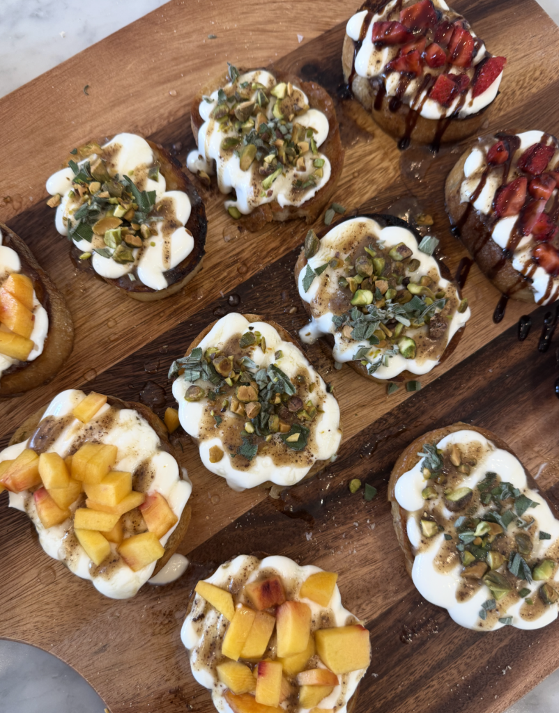 Nine pieces of toast topped with whipped ricotta, chopped herbs, pistachios, peaches, and strawberries with a drizzle of balsamic glaze and a hint of brown butter, all artfully arranged on a wooden board.